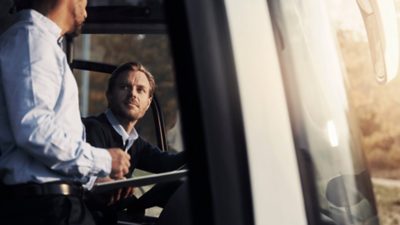 A male bus driver in the driver’s seat having a conversation with a man standing up.