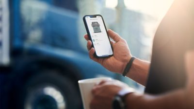 Närbild på en person som håller en kaffekopp i ena handen och en smartphone med My Truck-appen öppen i den andra