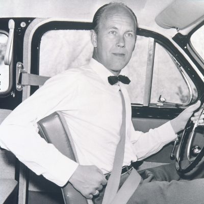 Nils Bohlin demonstriert den Dreipunkt-Sicherheitsgurt in einem Auto. Er trägt eine Fliege und hält den Gurt quer über der Brust.