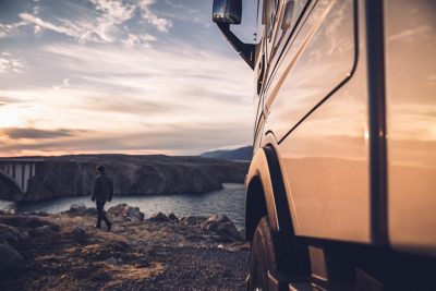 A driver sits on a rock looking out to sea at sunset, with his Volvo FH truck parked beside him