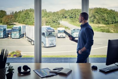 Asmuo, laikantis planšetinį kompiuterį, žiūri pro langą į Volvo Trucks, pastatytus kieme apačioje.