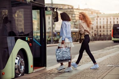 Two women about to board a full hybrid bus in a city location