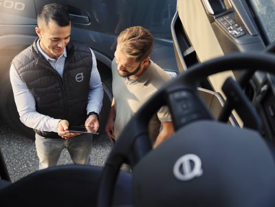 Dva dospělí muži stojí venku vedle nákladního vozu Volvo, jeden drží tablet. Snímek je pořízen z kabiny nákladního vozu a je na něm jasně vidět volant s jasně viditelným emblémem Volvo. Prostředí působí profesionálně, jeden muž má na sobě značkovou vestu a druhý je v neformálním oblečení. Na vnější straně nákladního vozu je viditelné číslo „500“, které označuje model. Osvětlení je přirozené, což evokuje denní pracovní nebo školicí scénu.