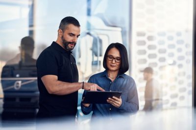 Dva lidé stojí vedle sebe v prodejně nákladních automobilů Volvo a dívají se na tablet. V pozadí je vidět rozostřený nákladní vůz a další personál.