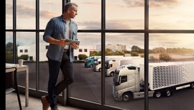 Vyras, laikantis skaitmeninį įrenginį, žiūri pro langą į sunkvežimių parką 
