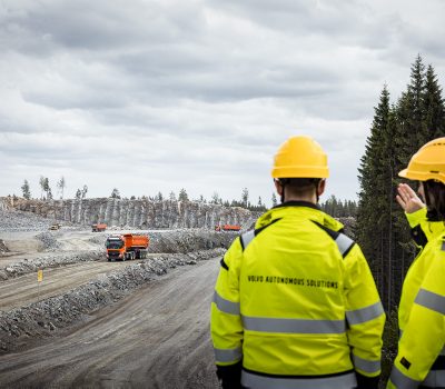 2 men are seen in the image, seemingly discussing the truck driving towards them from a far distance. There are 3 trucks that can be seen in the distance and all of this is inside a quarry. There is a row of pine trees in front of the workers who are at the right corner of the image.