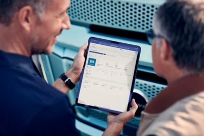 Two people stand beside a turquoise Volvo truck while one of them holds a tablet displaying fleet or vehicle‑performance data on the screen. The close-up view focuses on the tablet interface and the truck’s front grille in the background.