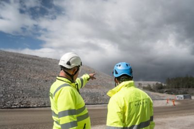 Two workers in high-visibility jackets and helmets stand at a construction or quarry site under a dramatic, cloudy sky. The person on the left points toward a rocky embankment, while the other, wearing a blue helmet, looks on. The jacket on the right has a visible Volvo logo. The ground is unpaved, with traffic cones and a small site office visible in the background near a tree line.