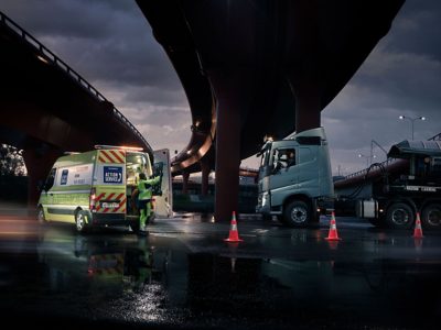 En Volvo Action Service-varevogn er parkeret ved siden af en lastbil under en motorvejsbro