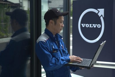 A mechanic in a blue jumpsuit stands by a glass wall, working on a laptop.