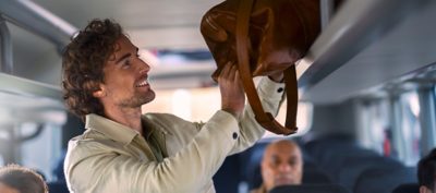 Hymyilevä linja-auton matkustaja laittamassa laukkua Volvo-sähkölinja-auton matkatavarahyllylle