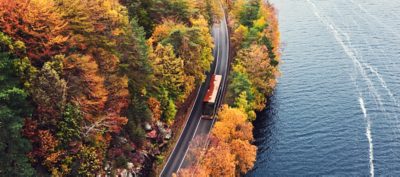A Volvo 9700 DD viewed from above driving on a narrow country lakeside road surrounded by a forest in autumn colours
