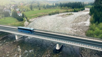 A Volvo 9700 crossing a bridge over a river with a village in the background.