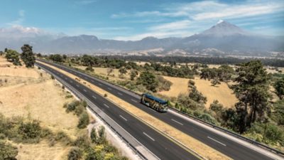 A Volvo 9800 6x2 driving on motorway through landscape with mountain view.