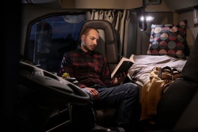 Kierowca ciężarówki Volvo odpoczywa w kabinie sypialnej, czytając książkę przy schludnie pościelonym łóżku w przytulnym, domowym wnętrzu.
