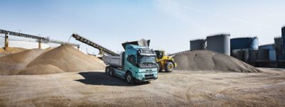 Volvo electric tipper truck with a turquoise cab is positioned in front of large gravel piles as a wheel loader deposits material into the raised tipper body. Conveyor structures, storage tanks, and industrial equipment surround the scene, indicating active operations at an aggregate or quarry processing site.