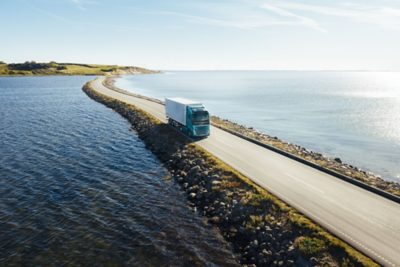 Un camión eléctrico Volvo, con cabina turquesa y remolque de caja blanca, avanza por una estrecha carretera de la costa flanqueada por rocas, con el mar abierto a ambos lados bajo la luz del día. La escena está captada desde un ángulo elevado, lo que resalta el movimiento del camión a través del pintoresco paisaje costero.