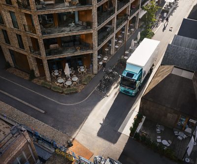 Luchtfoto van een elektrische Volvo-vrachtwagen met een turquoise cabine en een witte laadbak, rijdend door een smalle straat tussen moderne woongebouwen. Buiten zitjes, fietsen en architectonische wandelpaden vormen het decor en laten zien hoe het voertuig zich door een compacte stedelijke omgeving beweegt.
