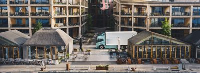 Een Volvo FH Aero Electric vrachtwagen met een turquoise cabine en een lange oplegger rijdt over een kronkelende bergweg langs rotsachtige hellingen, met open water en in de verte heuvels zichtbaar bij helder daglicht. De scène is vastgelegd vanuit een frontaal driekwart perspectief, waardoor de beweging van het voertuig door het landschap wordt benadrukt.