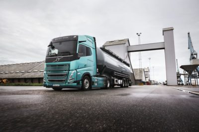 Un camión eléctrico Volvo FH Electric, con cabina turquesa y remolque cisterna oscuro, circula por una planta industrial; avanza entre grandes estructuras grises y depósitos sobre una superficie pavimentada y húmeda. La escena está capturada desde un ángulo bajo de tres cuartos frontal, lo que resalta el movimiento del vehículo y su diseño aerodinámico.