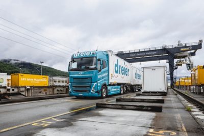 Volvo truck with a turquoise cab and branded box trailer drives through an intermodal terminal area, surrounded by rail equipment, overhead wires, and freight infrastructure under overcast skies. A large terminal sign and surrounding containers and platforms are visible in the background.