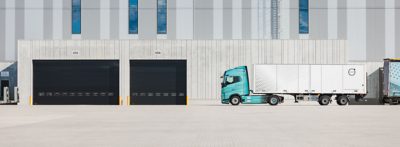 Un camión eléctrico Volvo, con cabina turquesa y remolque tipo caja, permanece estacionado frente a una amplia zona de carga industrial, delante de altos muros de hormigón con dos puertas basculantes negras numeradas 655 y 656. La imagen muestra el vehículo detenido en un patio pavimentado adyacente a un almacén.