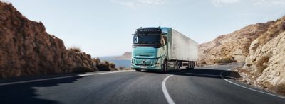 Een Volvo FH Aero Electric vrachtwagen met een turquoise cabine en een lange oplegger rijdt over een kronkelende bergweg langs rotsachtige hellingen, met open water en in de verte heuvels zichtbaar bij helder daglicht. De scène is vastgelegd vanuit een frontaal driekwart perspectief, waardoor de beweging van het voertuig door het landschap wordt benadrukt.