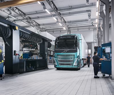 Un camión eléctrico Volvo está estacionado en una estación de carga cubierta en una noche lluviosa, con el pavimento mojado reflejando las luces de la estructura. La escena incluye dos unidades de carga y una zona boscosa al fondo.