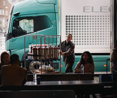 Elektrische Volvo-vrachtwagen staat 's nachts geparkeerd onder een felverlicht laadplatform, waarbij de vrachtwagen wordt omlijst door bovenliggende balken en lineaire ledverlichting.