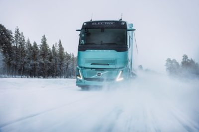 Een elektrische Volvo-truck rijdt met hoge snelheid over een besneeuwde weg in winterse omstandigheden en jaagt daarbij een wolk fijne sneeuw op. Op de achtergrond, bij helder daglicht, staan ​​met sneeuw bedekte bomen, wat de testomgeving in koude weersomstandigheden benadrukt.