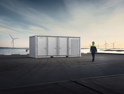 Image of a Volvo PU2000 on a road with a body of water in the background. The background contains one windmill to the left in the distance, while towards the right there are four windmills. All those 4 windmills are increasingly further away from the point of view of the camera. In the foreground there is a lady walking candidly next to the PU2000. In front of the PU2000 is just asphalt, part of a paved road. 