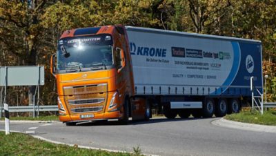 Orangefarbener Volvo FH Globetrotter-Lkw mit Krone-Anhänger fährt auf einer kurvigen Straße, umgeben von herbstlichen Bäumen.