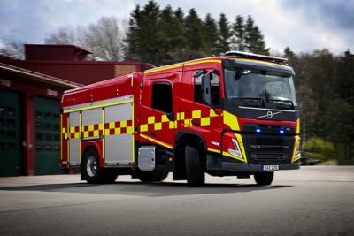 Czerwony wóz strażacki Volvo z żółtymi szachownicowymi oznaczeniami odjeżdża od remizy strażackiej, mając włączone z przodu niebieskie światła ostrzegawcze.