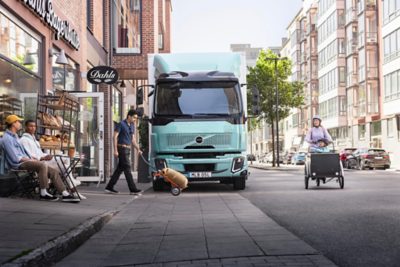 Un veicolo per le consegne Volvo FL Electric parcheggiato fuori da un panificio cittadino mentre un corriere scarica la merce e un genitore passa in bicicletta con un bambino su una cargo bike.