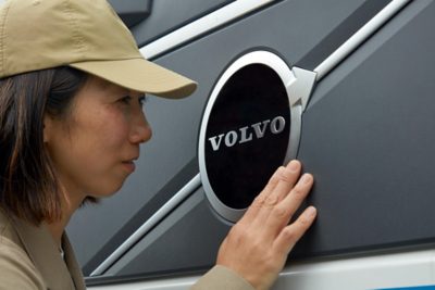 A person wearing a tan cap and jacket is closely examining and touching a large Volvo emblem.