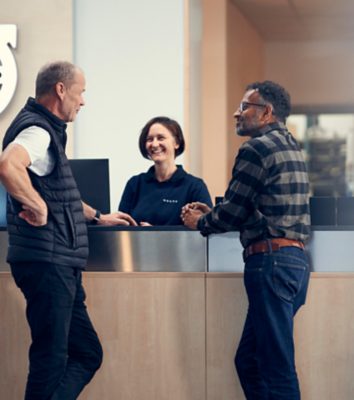 Dwóch klientów rozmawia z uśmiechniętym przedstawicielem serwisu Volvo przy recepcji salonu dealerskiego.
