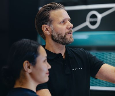 Twee mensen staan ​​in een werkplaats of servicegebied. Een van hen draagt ​​een zwart Volvo-shirt en wijst naar een turquoise vrachtwagengrille, waarop het Volvo-embleem op de achtergrond zichtbaar is. De scène draait om een ​​gezamenlijke discussie in een professionele omgeving.