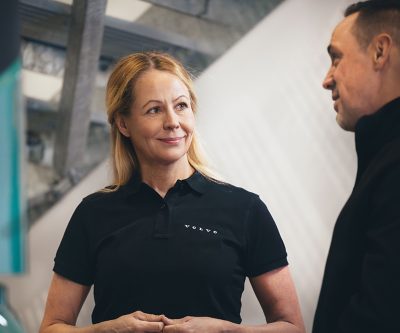 Two people stand indoors in a Volvo facility, with one wearing a dark Volvo-branded shirt while they engage in conversation. The background shows a modern industrial or office setting with light-colored structural elements.
