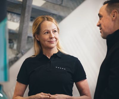 Twee mensen staan ​​binnen in een Volvo-fabriek, waarvan één een donker shirt met het Volvo-logo draagt, terwijl ze een gesprek voeren. De achtergrond toont een moderne industriële of kantooromgeving met lichtgekleurde bouwelementen.