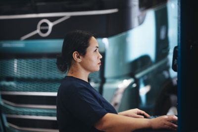 A person wearing a dark shirt is positioned at a workstation inside a service area, operating equipment while a turquoise Volvo truck is parked in the background. The scene highlights an indoor workshop environment with the truck’s front grille and cab visible behind the person.