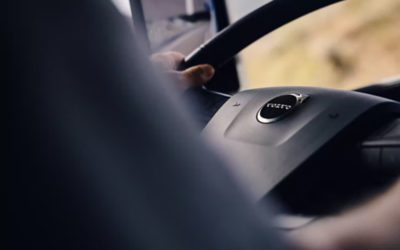Close-up of a driver’s hands on a Volvo truck steering wheel while driving, with the road visible ahead through the windshield.
