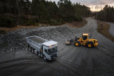 Samochód ciężarowy Volvo załadowany żwirem stoi na szerokiej żwirowej powierzchni obok żółtej ładowarki kołowej Volvo w miejscu przypominającym kamieniołom, gdzie znajdują się duże stosy kamieni.