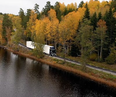 Volvo kamion sandučar u vožnji uskim putem pored jezera, okružen jesenjim drvećem u šumskom pejzažu, snimljen iz zraka.