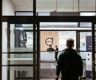 Uitzicht door een glazen deuropening op een receptiebalie in een Volvo-vestiging, waar twee medewerkers achter een bureau staan ​​onder een groot rond Volvo-logo op een houten wandpaneel. Op de voorgrond is een persoon te zien die het gebouw binnengaat, en links is een deel van een vrachtwagen zichtbaar in de werkplaats.
