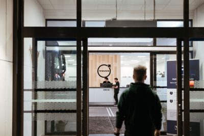 View through a glass entrance toward a reception counter inside a Volvo facility, where two staff members stand behind a desk beneath a large Volvo circular symbol on a wooden wall panel. A person is entering the building in the foreground, and part of a truck is visible inside the workshop area to the left.