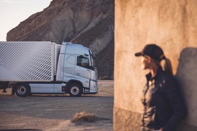 Nákladní automobil Volvo se skříňovým návěsem stojí zaparkovaný v kamenité pouštní krajině a v popředí se rozostřená osoba v čepici a bundě opírá o zeď.