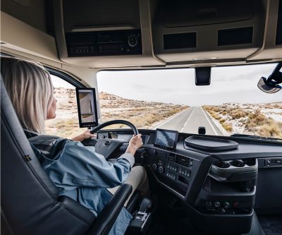 Vozač kamiona sjedi u kabini Volvo kamiona i vozi otvorenim putem kroz suhi pejzaž, pogled iz kabine, vide se prikazi na instrument tabli i ekran bočne kamere.
