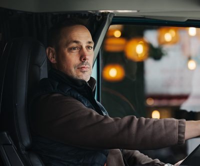 De persoon achter het stuur van een Volvo-vrachtwagen houdt het stuur met één hand vast, terwijl het warme licht van gebouwen of straatverlichting door het zijraam zichtbaar is. De afbeelding focust op de rijhouding en de interieurruimte van de cabine.