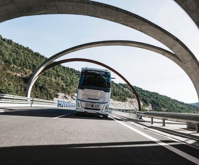 Volvo kamion se vozi autoputem koji prolazi ispod velikih zakrivljenih mostnih konstrukcija u planinskom krajoliku, snimljen iz niskog prednjeg ugla.