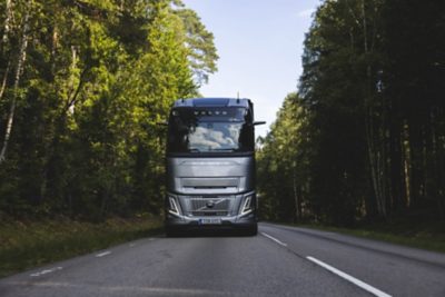 Front view of a Volvo truck driving along a two-lane road surrounded by dense green forest in daylight.
